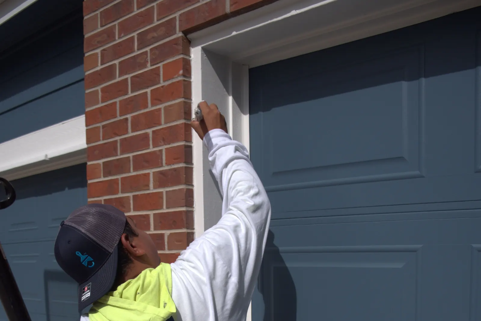 Kind Home painter cutting in white trim around a garage door on a brick exterior